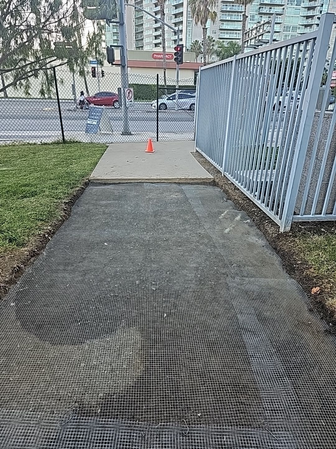 Pathway with textured surface, leading to a concrete walkway, bordered by grass, a fence, and a street.