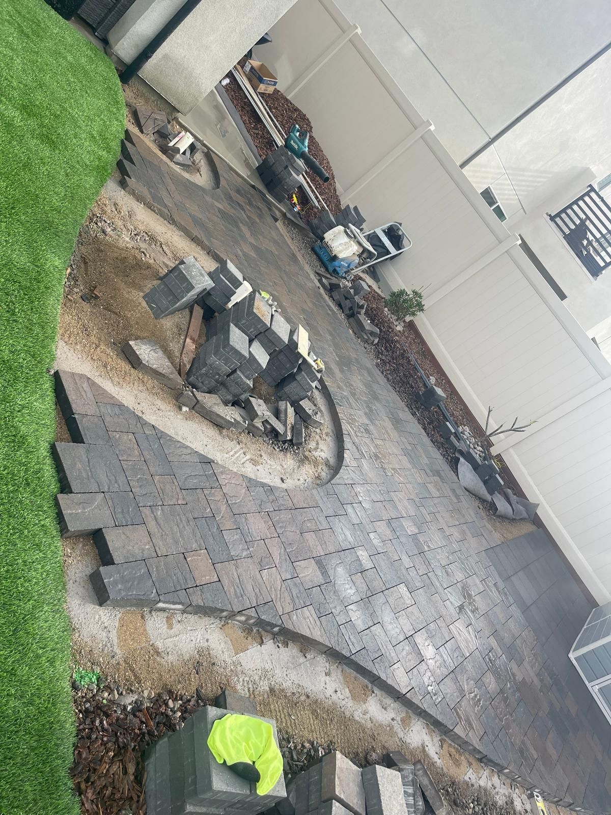 Backyard patio under construction with pavers, tools, and a white fence.