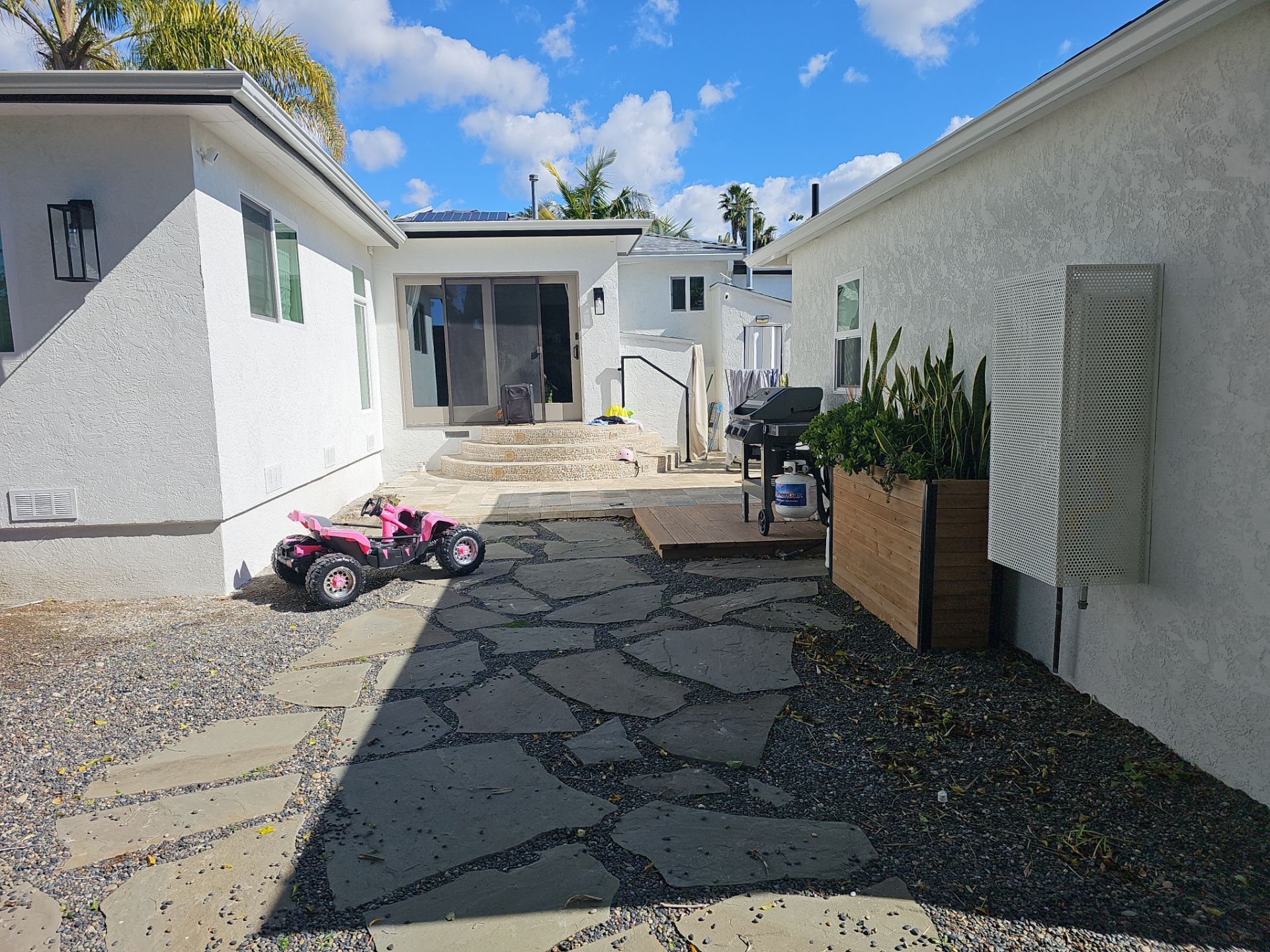 Stone pathway in a backyard with white stucco buildings. A child's pink ATV sits on the path.