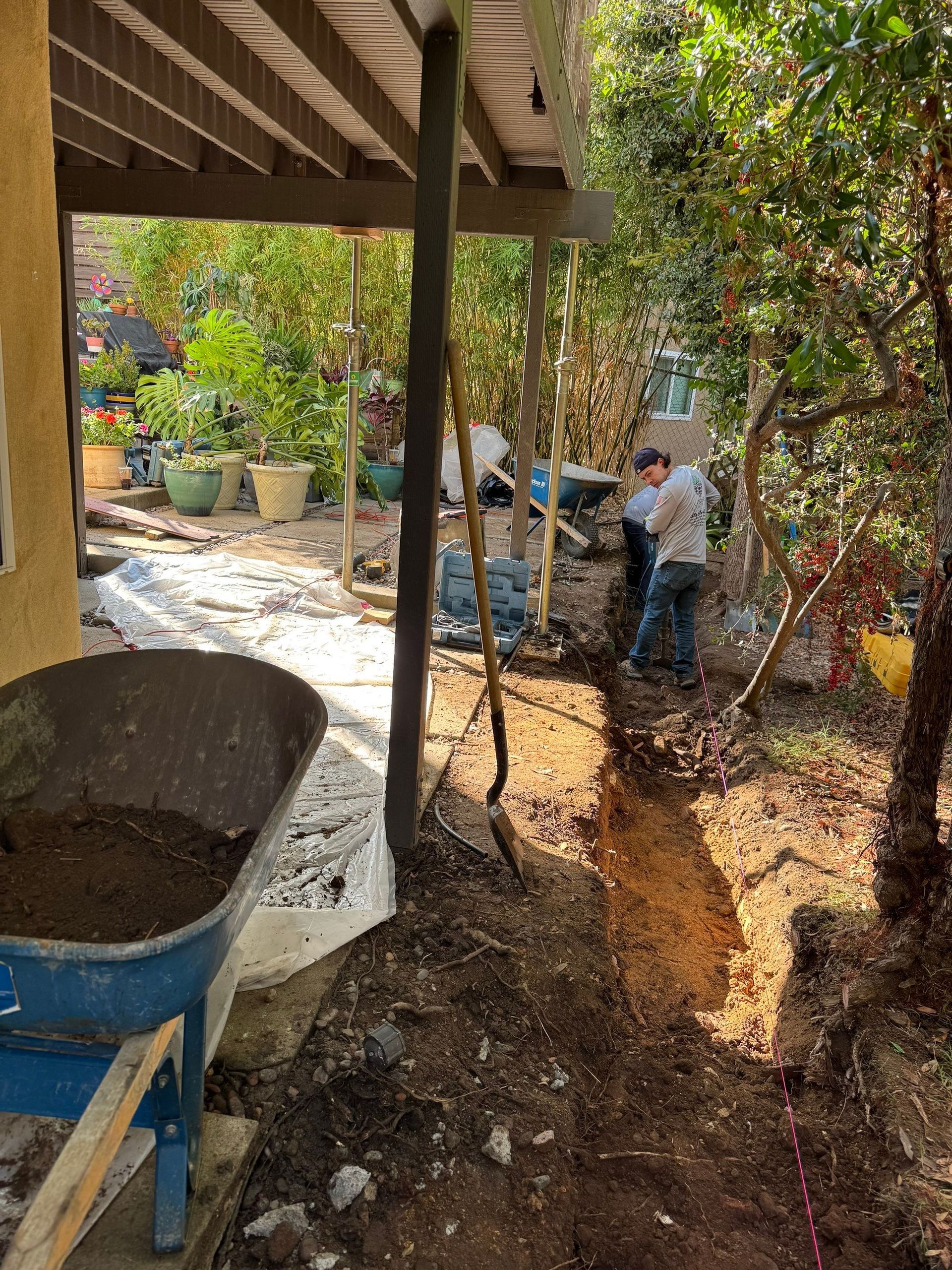 Construction work in a garden: two men dig a trench near a wooden deck, next to a wheelbarrow.