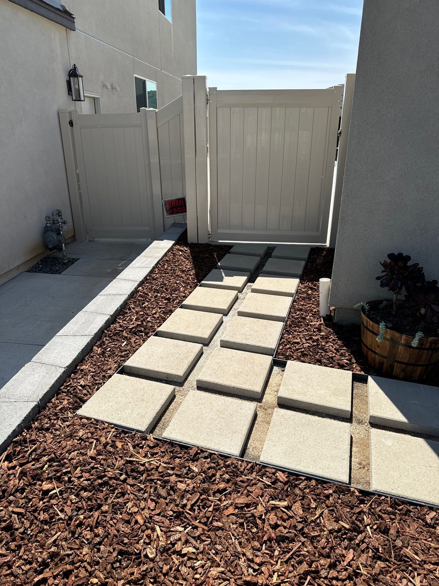 Pathway of square stepping stones through wood chips to a white gate.