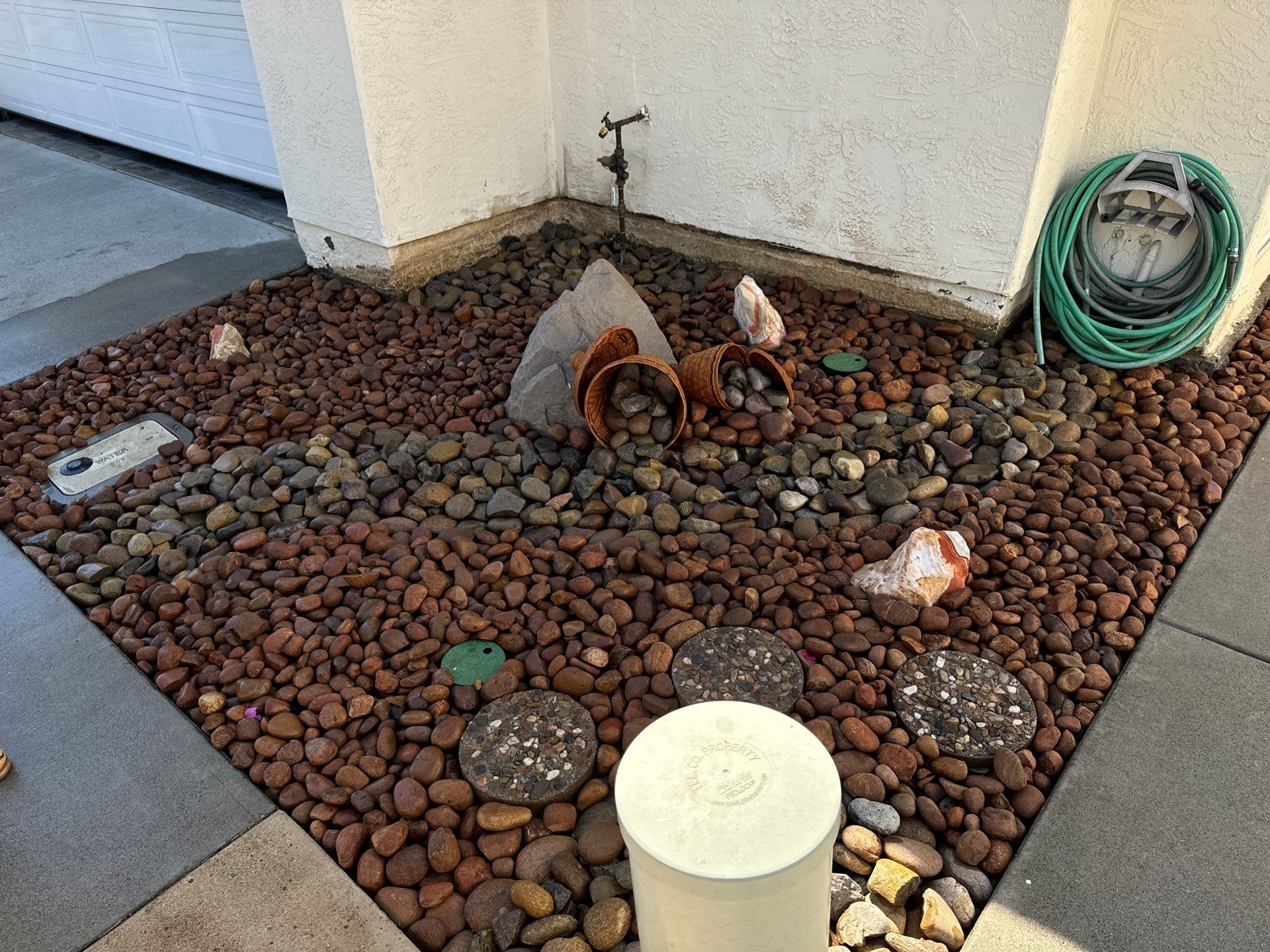 Rock-filled corner garden bed with decorative stones, pottery, and a coiled green hose next to a white wall.