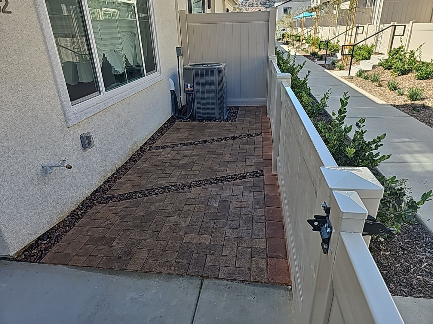 Small paved patio next to a house with an AC unit and a white fence, edged with brick.