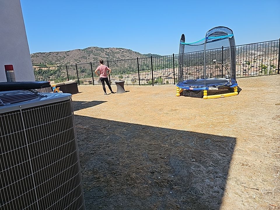Person walking on a gravel yard with a trampoline, air conditioning unit, and view of hills.
