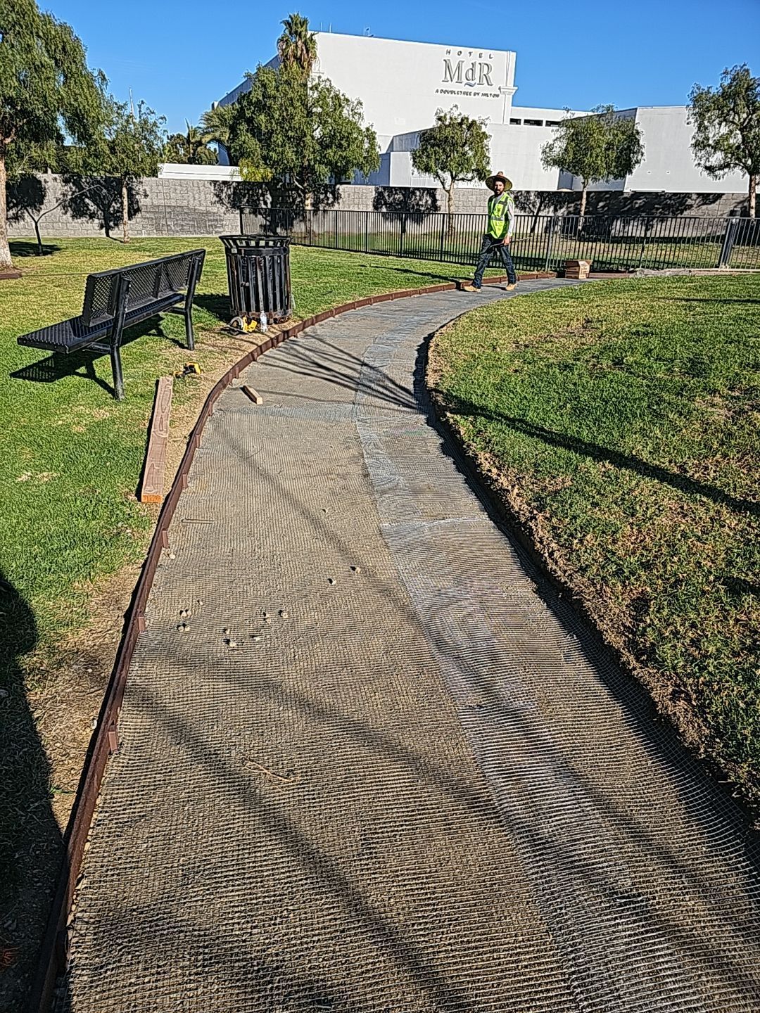 A park path curves through grass, with a bench on the left. A person is working in the background.