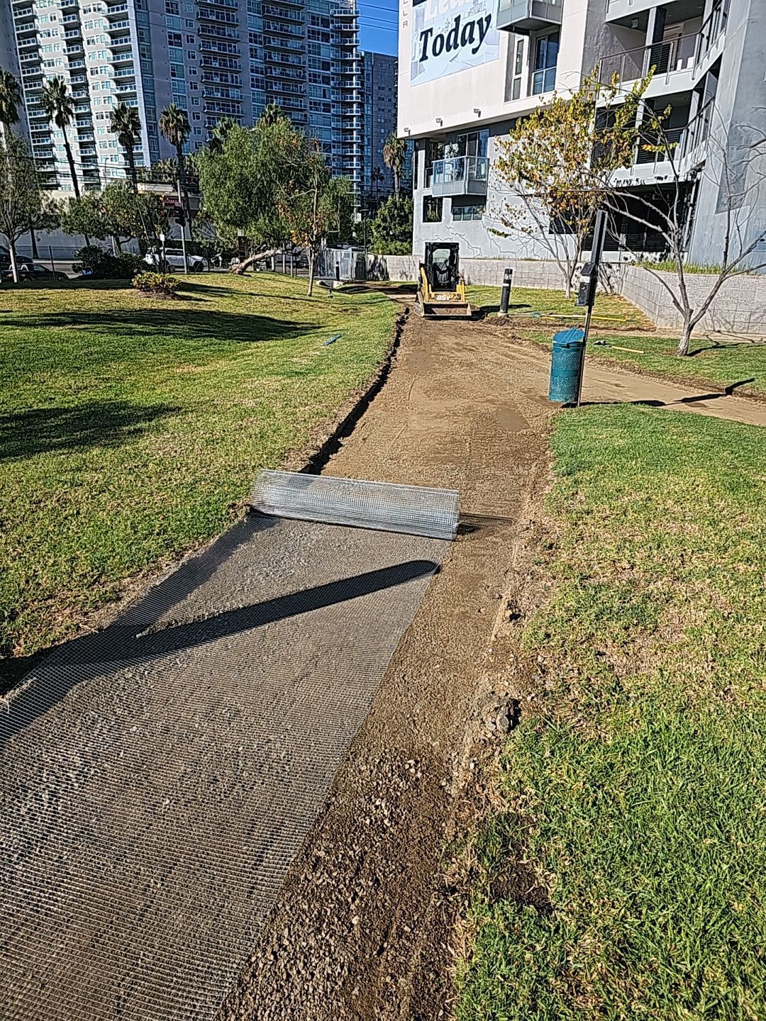 Path construction in a park: soil excavation, rolled mesh, and a building in the background.