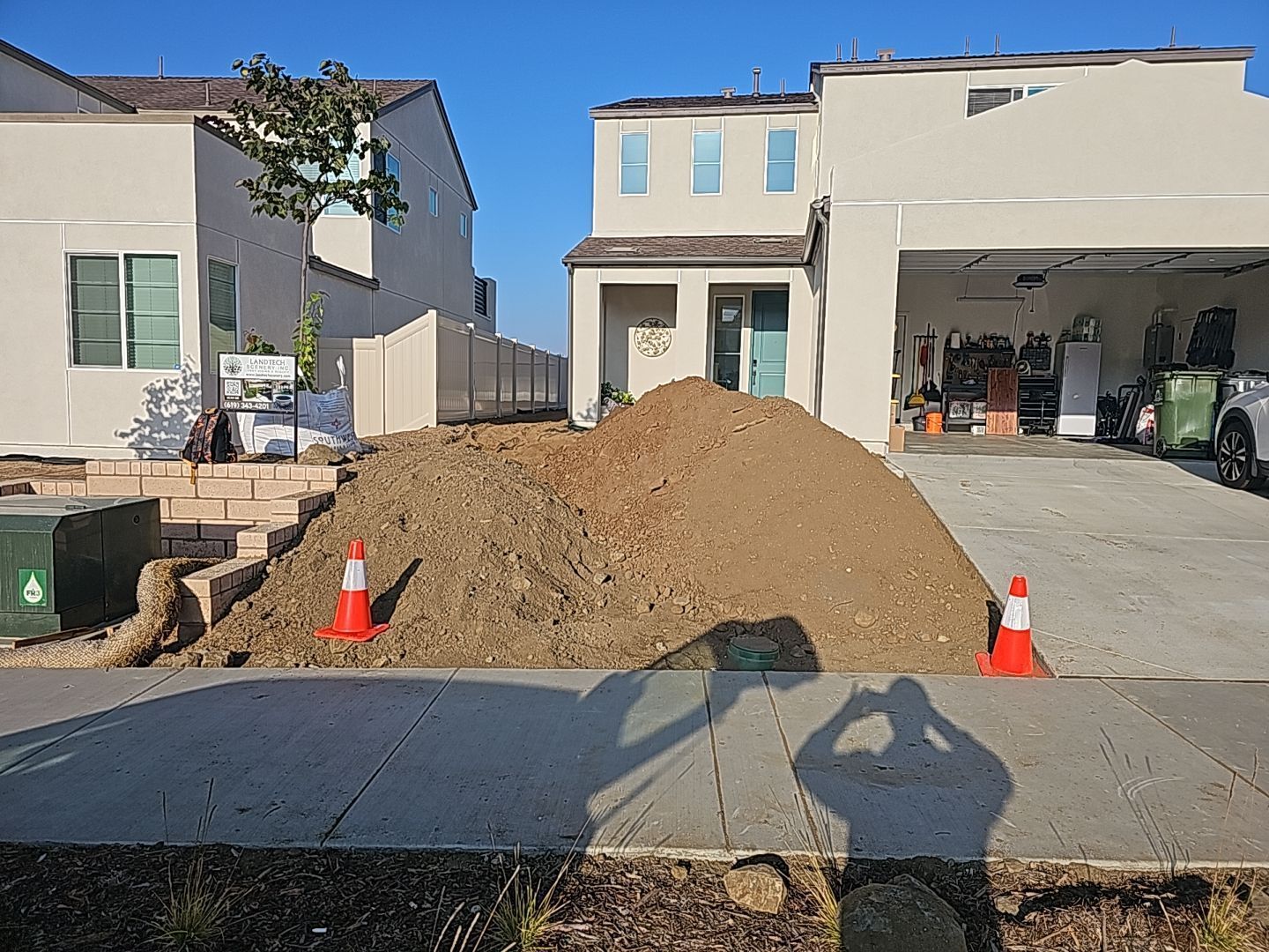 Pile of dirt in front of two-story houses; orange traffic cones; shadows cast on the sidewalk.