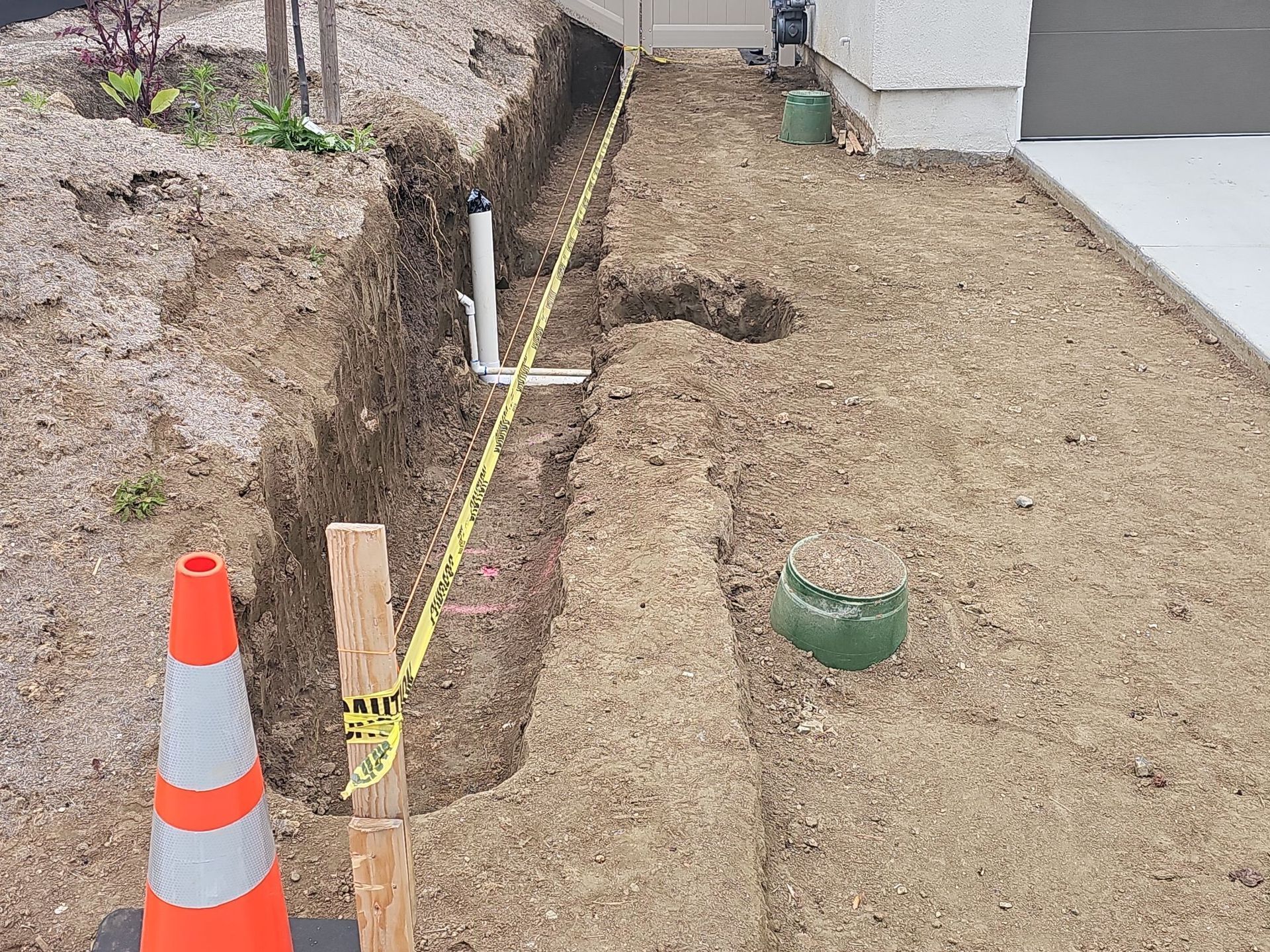 Trench dug in soil near a building, with caution tape and buried pipes. An orange traffic cone is visible.
