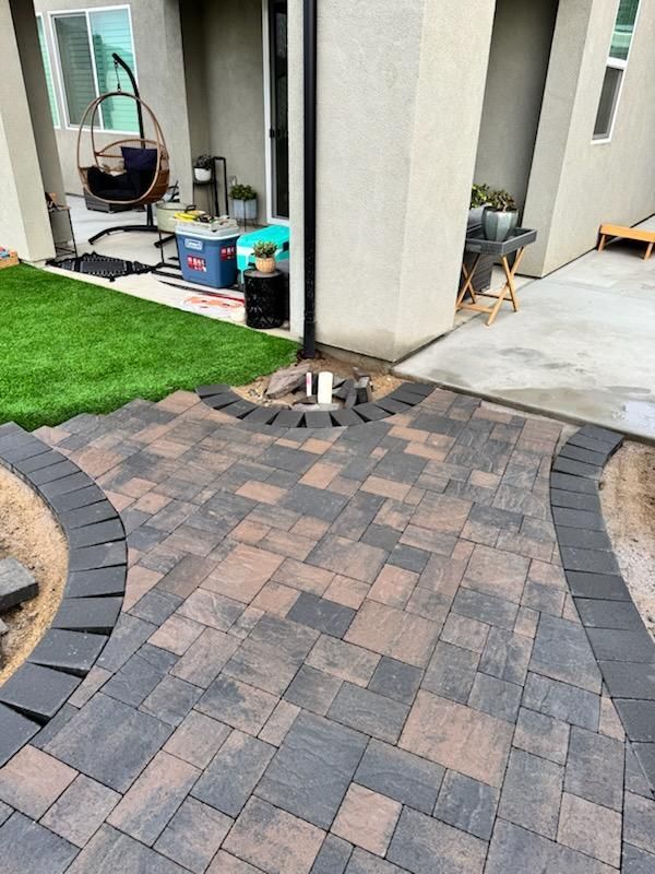 Patio with patterned pavers and curved border, adjacent to a house with a green lawn.