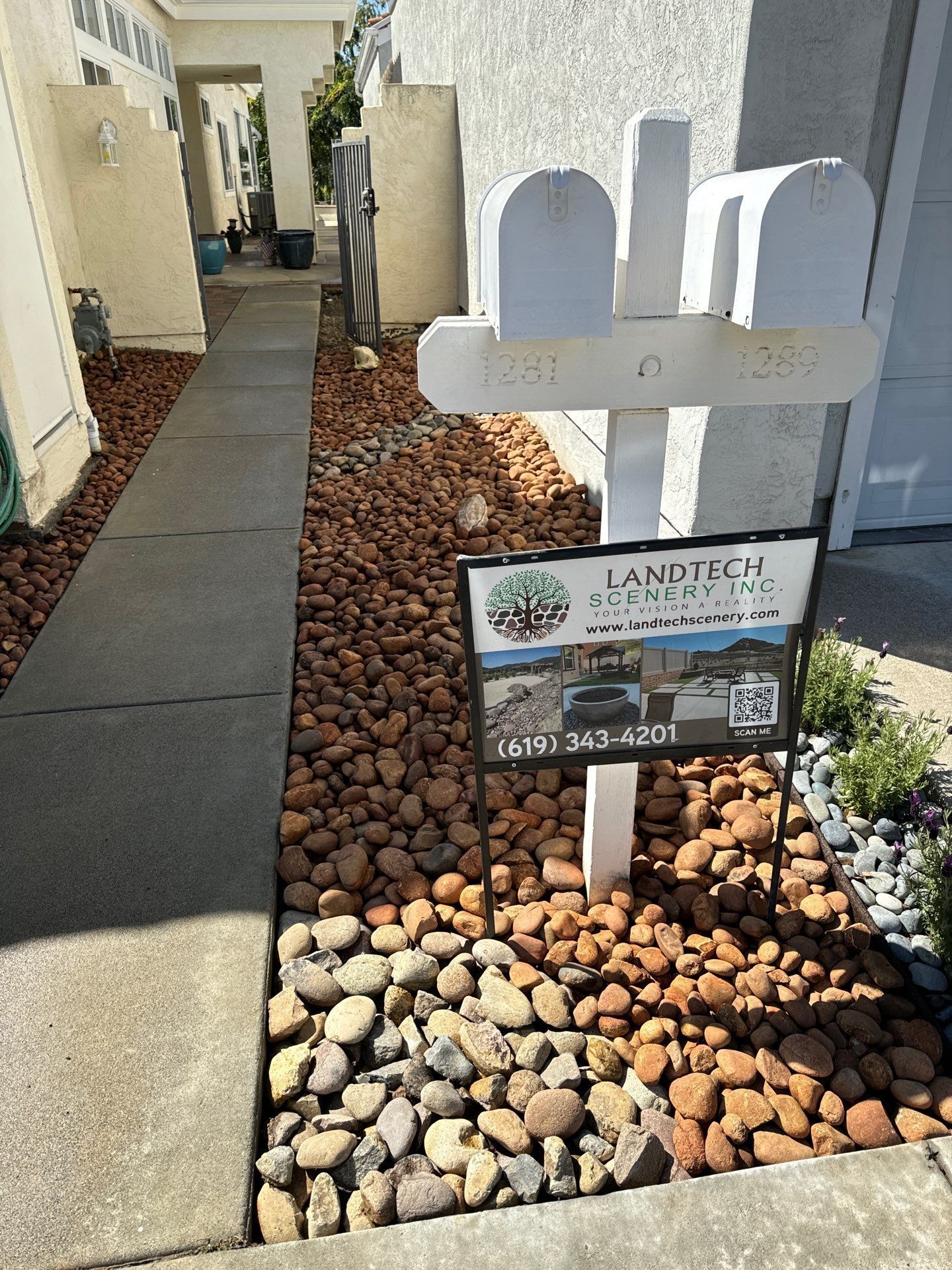 Mailboxes with a landscaping sign next to a walkway, surrounded by brown and gray rocks.