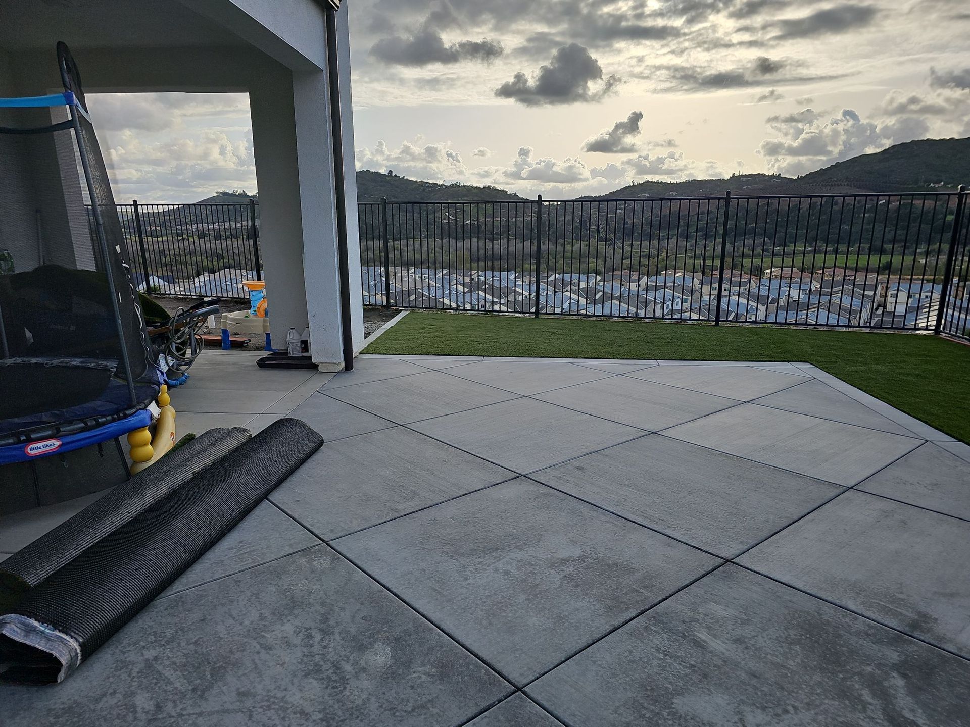 Patio with gray tiles, black railing, and distant houses with mountains under a cloudy sky.