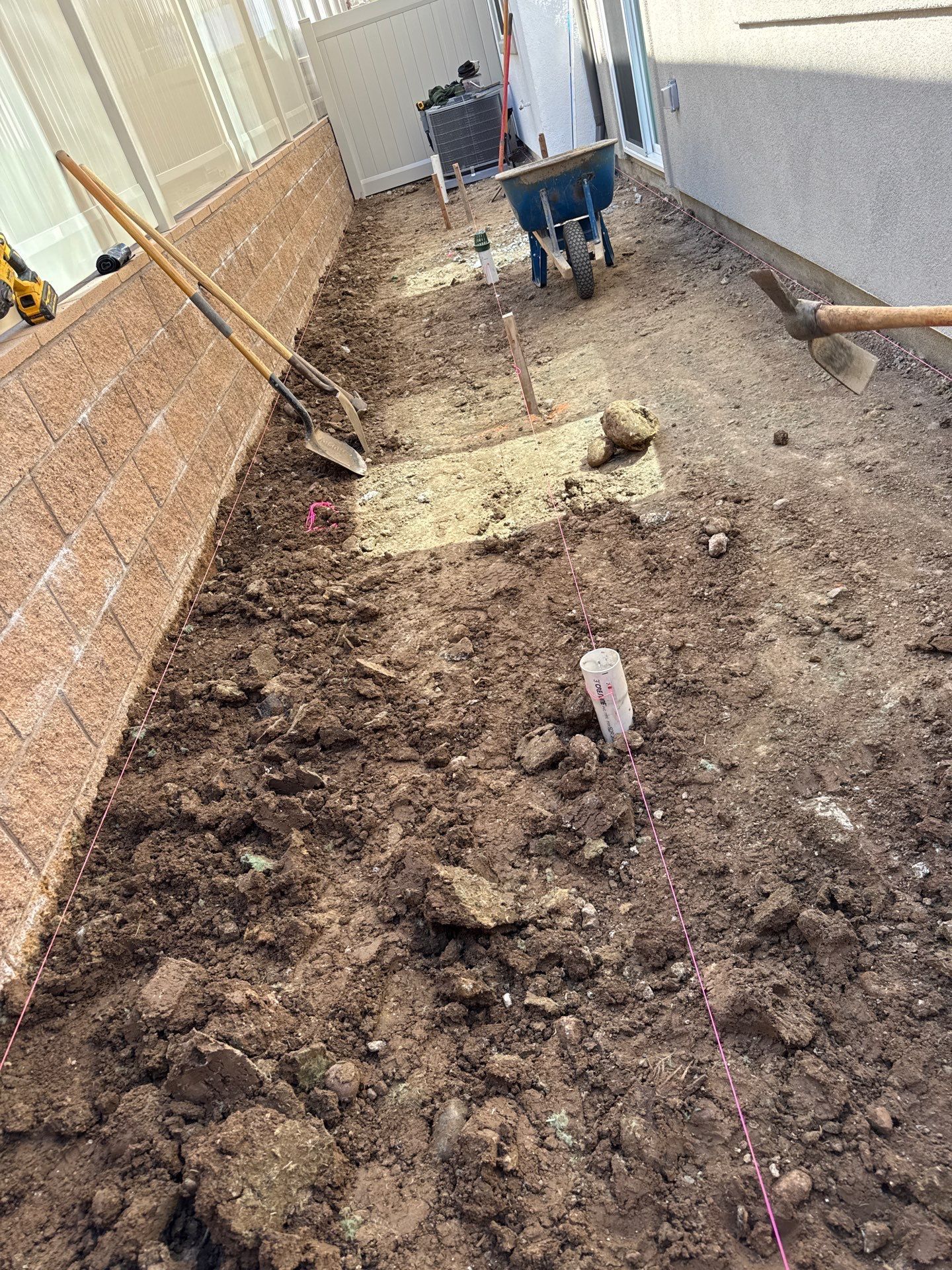 A narrow, excavated outdoor area with gardening tools and a wheelbarrow; construction site.