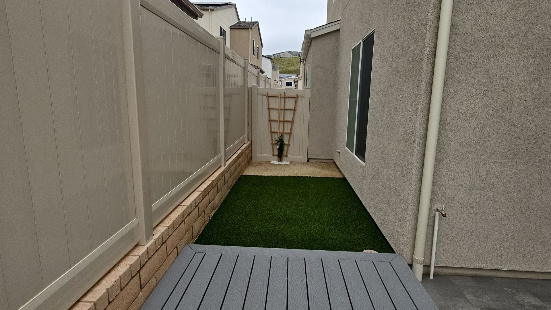 Narrow side yard with artificial turf, gray deck, beige fences, and a door at the end.