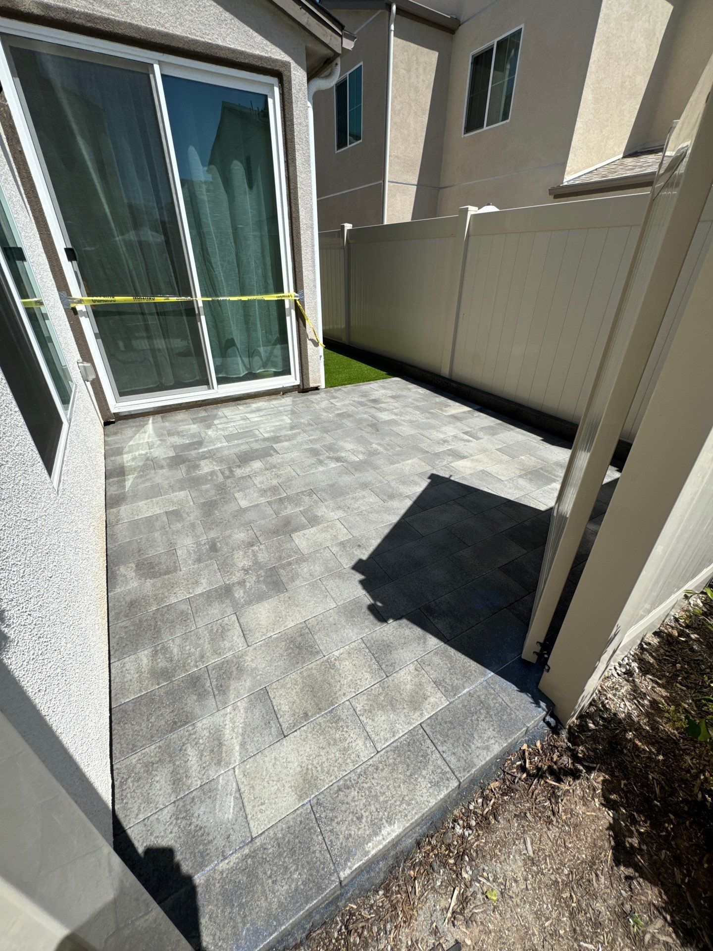 A paved patio with a sliding glass door. Gray pavers, white fence, and small patch of green grass.