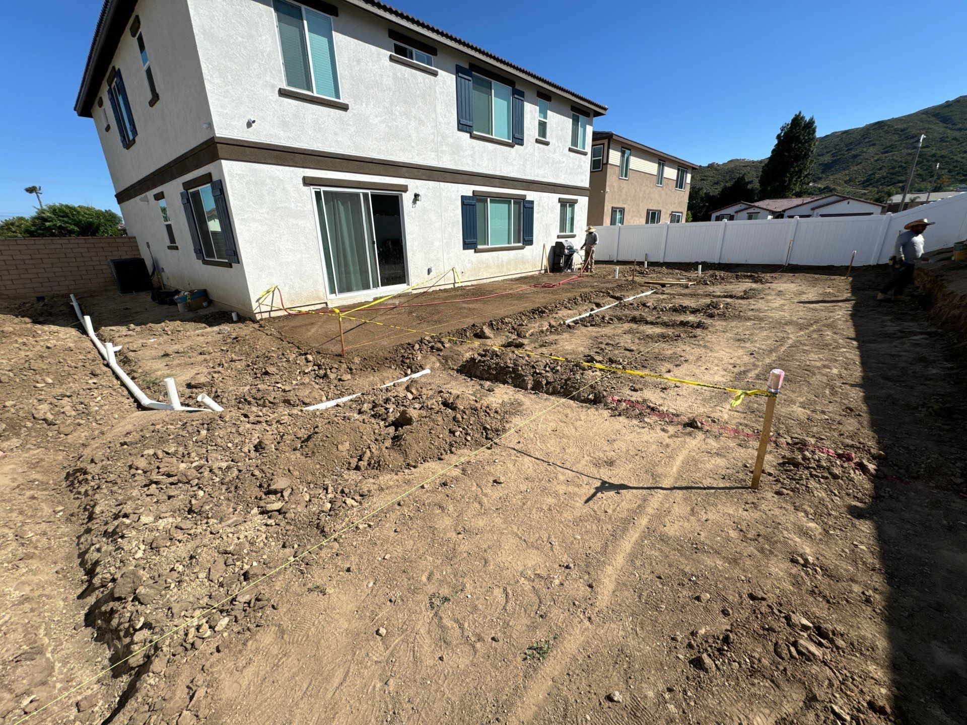 Backyard construction site: dug trenches, white house, clear sky.