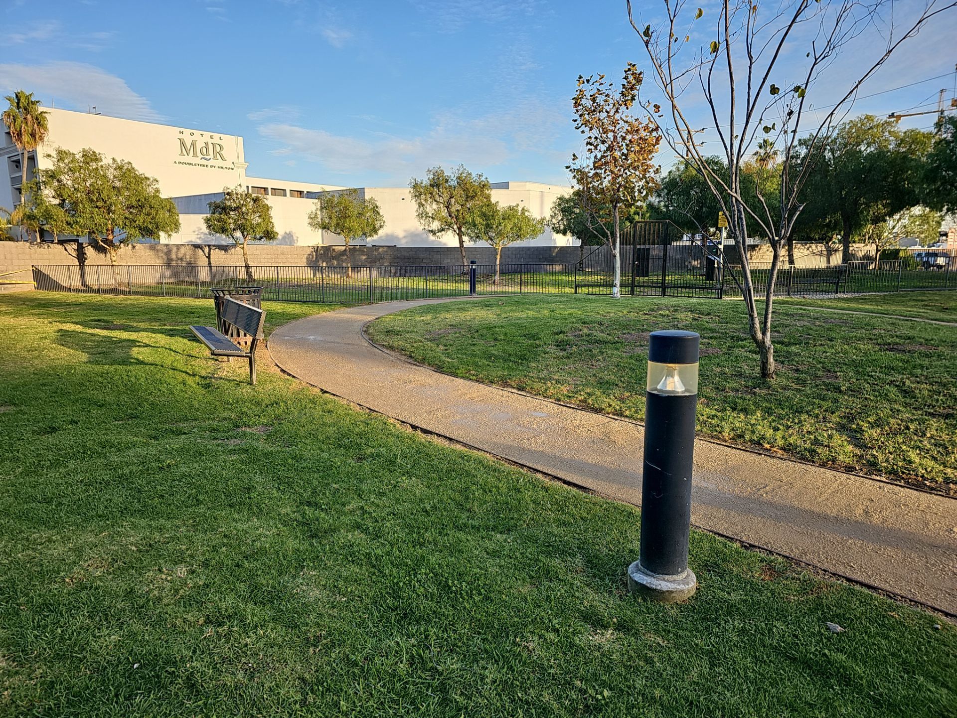 Park scene with a winding path, benches, trees, and a building in the background on a sunny day.