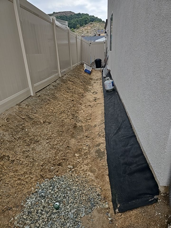 A narrow dirt yard between a beige fence and a white wall, partially covered with black fabric and gravel.