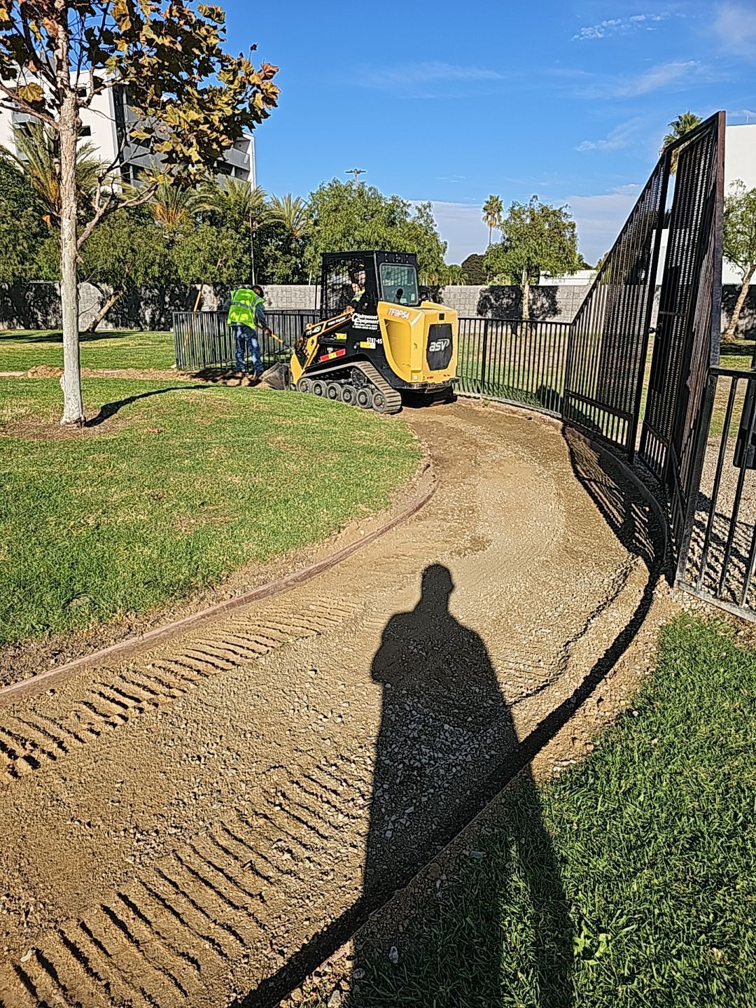 A small bulldozer grading a gravel path near a fence. A person in a green vest stands nearby, under a blue sky.