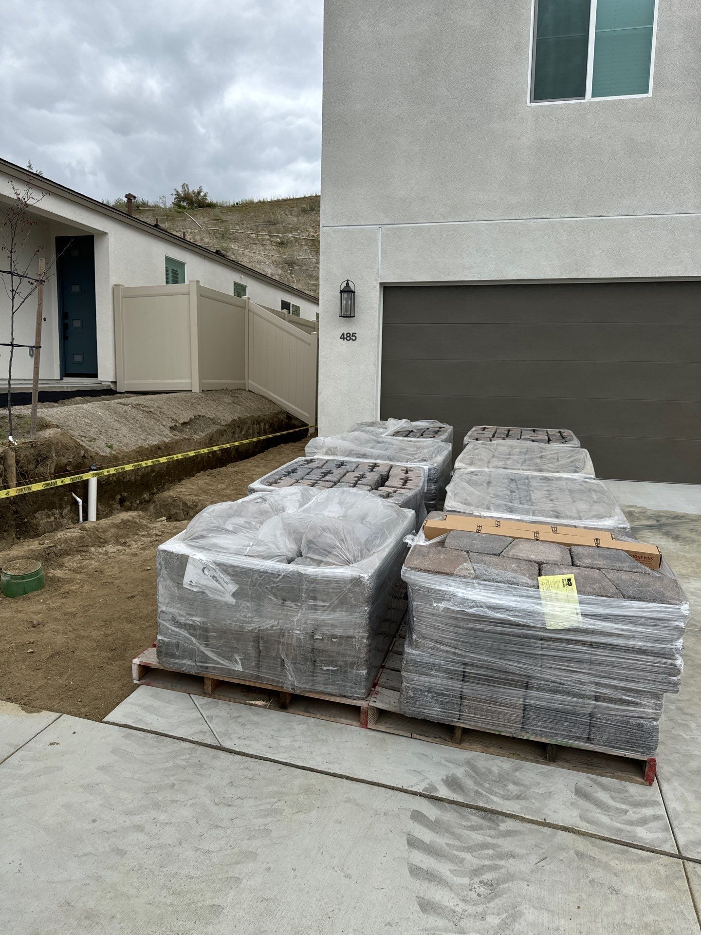 Pallets of paving stones sit on a driveway beside a house with a brown garage door.
