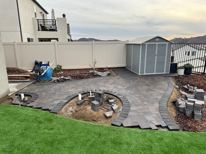 Backyard patio under construction with gray pavers, shed, and retaining wall.