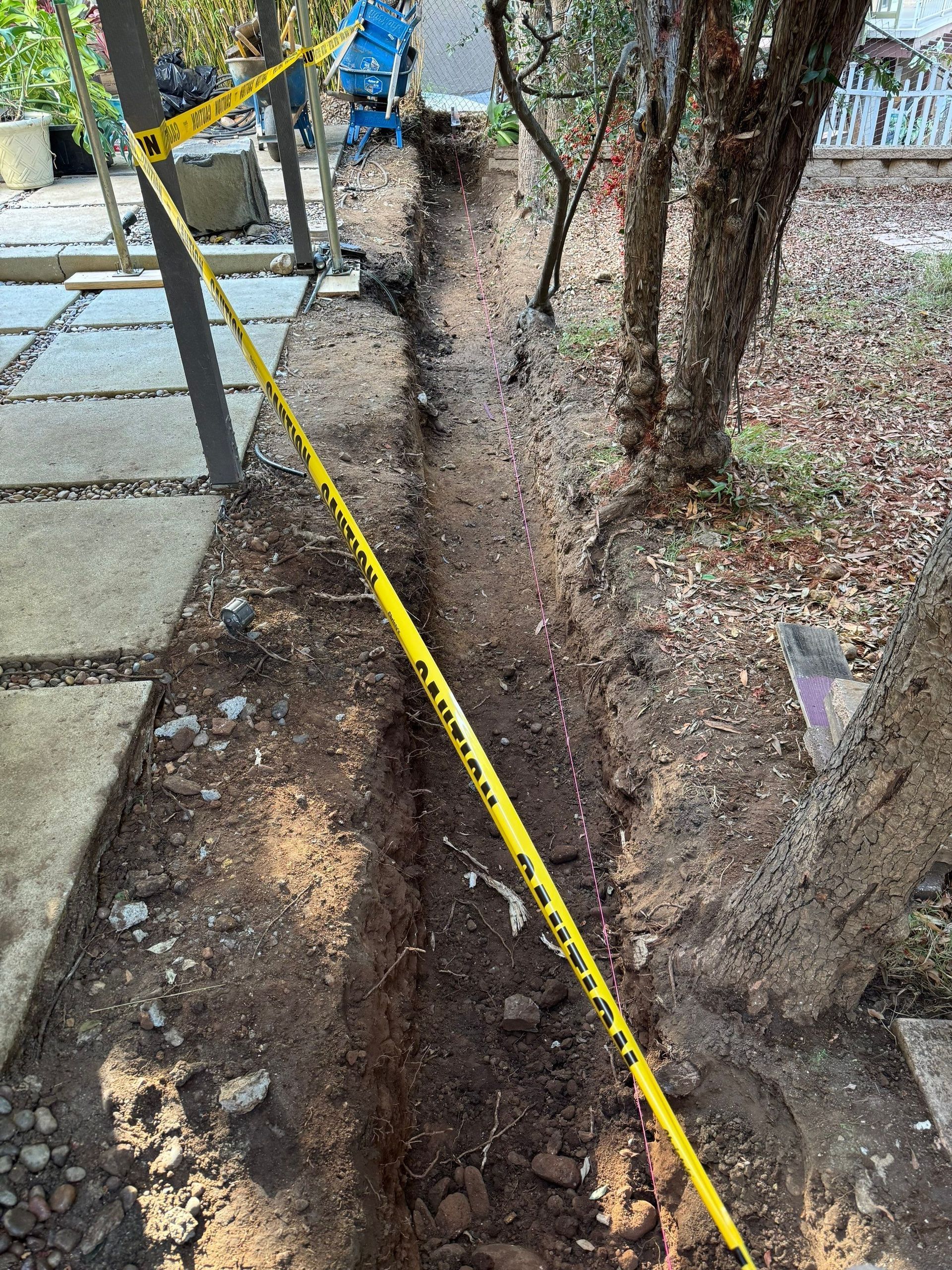 Long trench in dirt, marked with yellow caution tape, next to a paved walkway and trees.