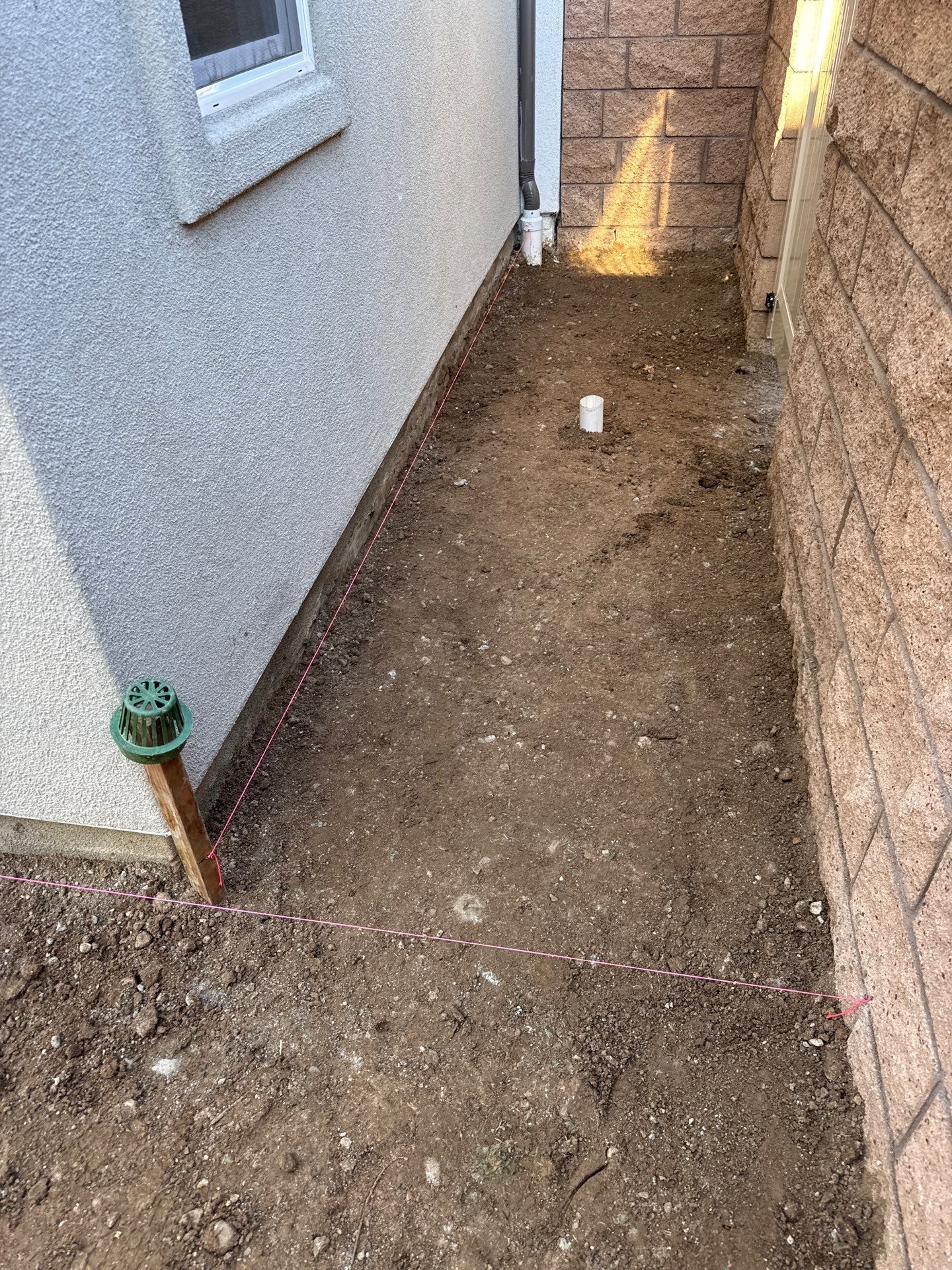 Narrow dirt pathway between a textured stucco wall and a brick wall, with plumbing visible.