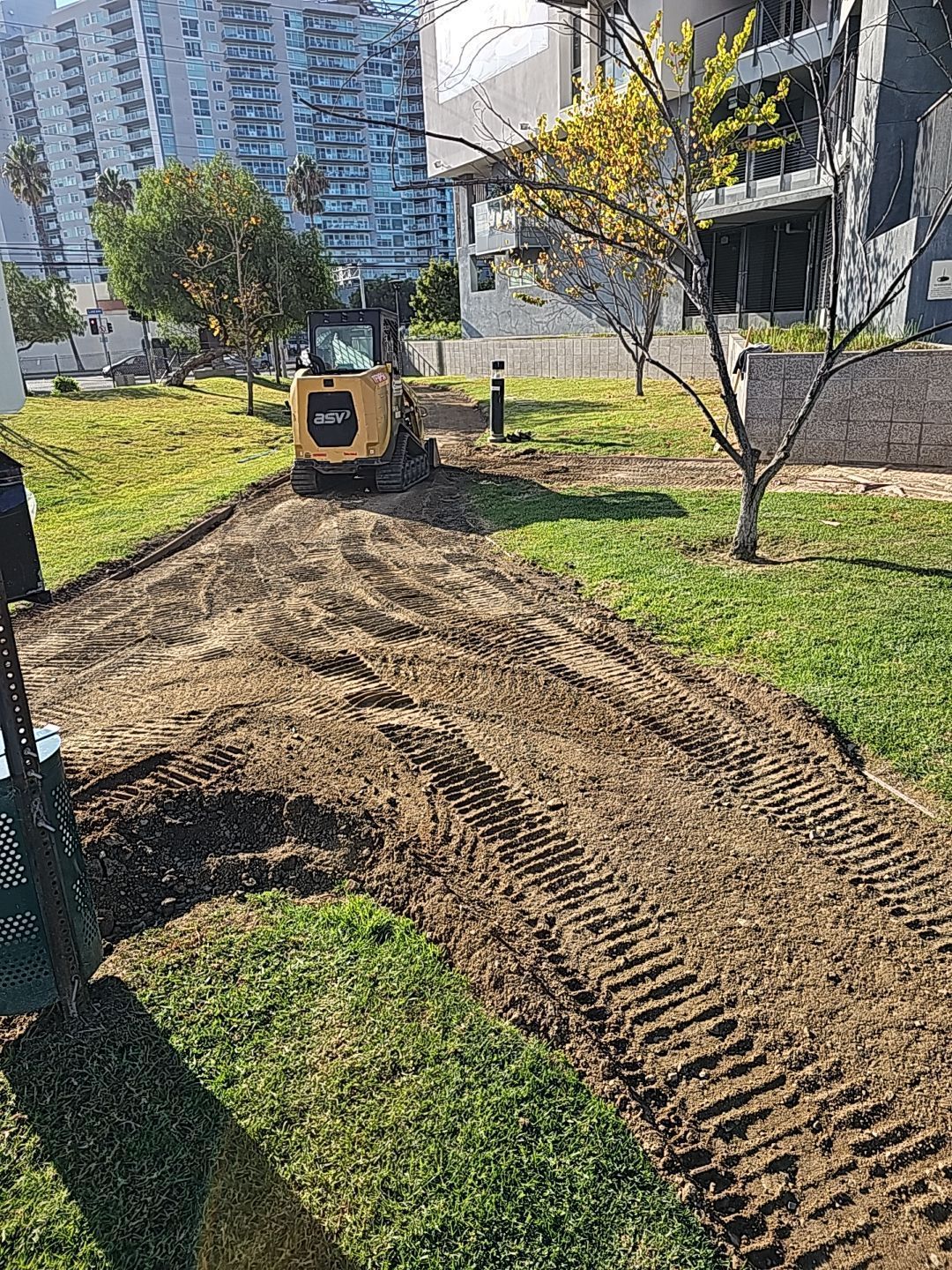 A yellow skid steer on a dirt path in a grassy area, preparing the ground. Buildings in background.