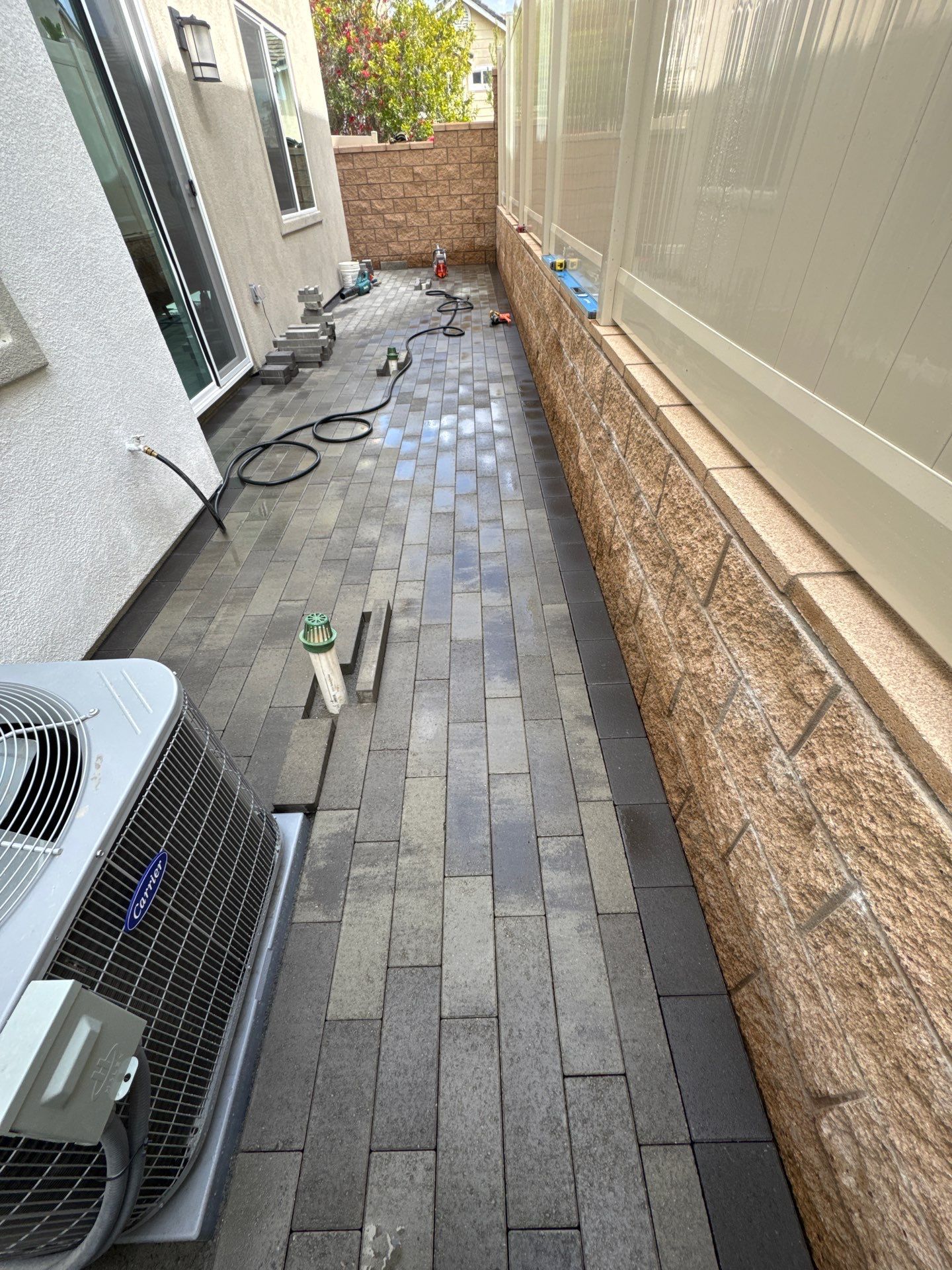 Brick pathway between a building and a decorative stone wall, near an AC unit.