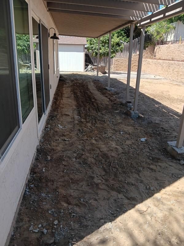 Dirt path along a building with sliding glass doors, under a pergola structure.