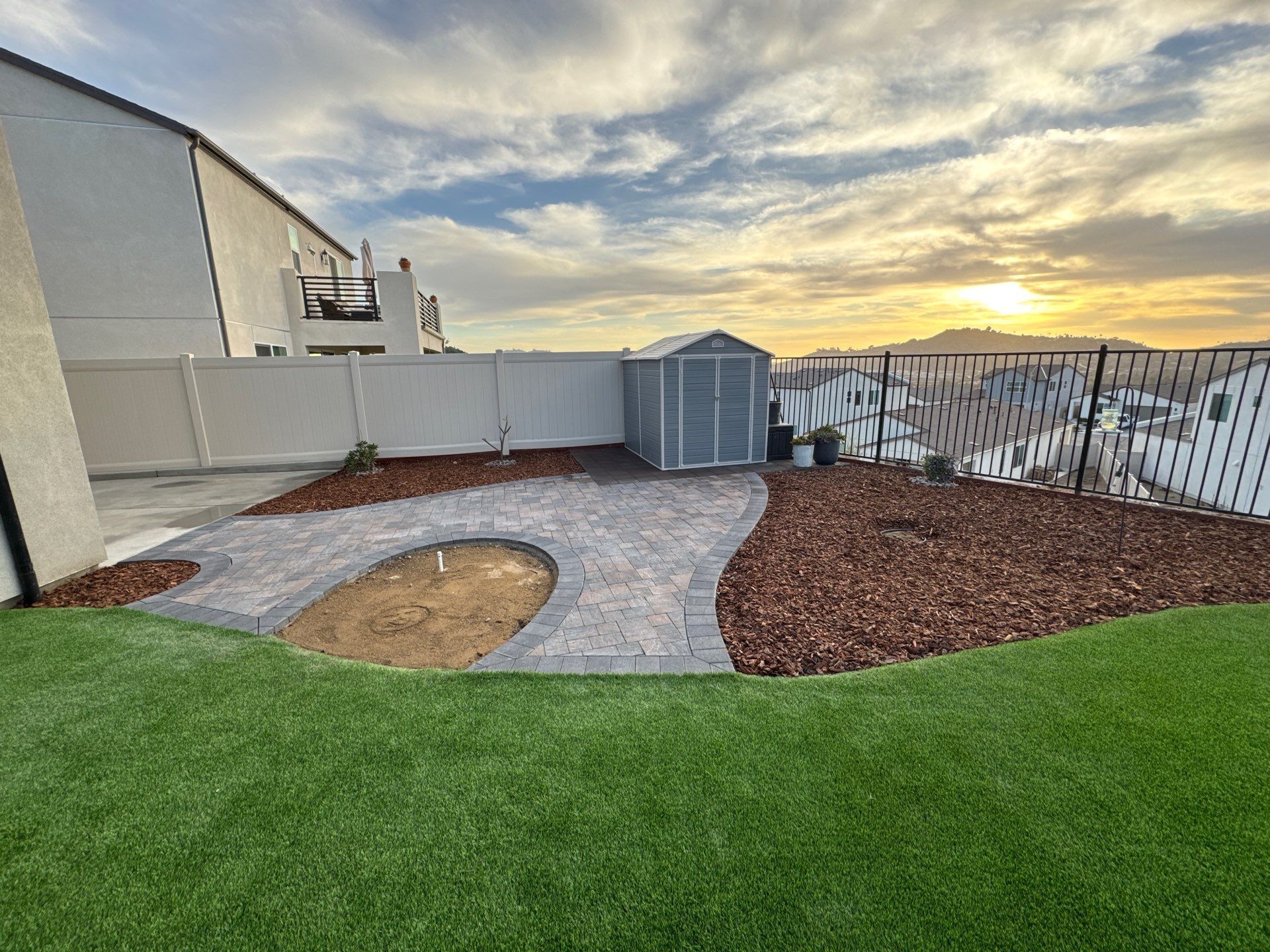 Backyard with shed, stone path, mulch, artificial turf, and fence against a sunset.