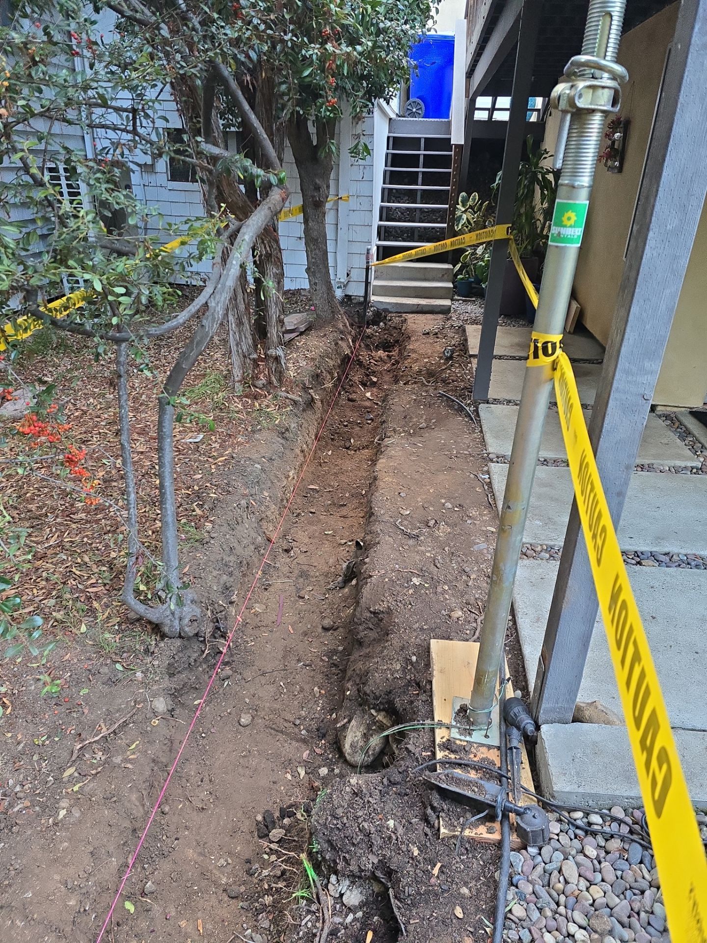 A trench dug in dirt, yellow caution tape, and support post next to stairs and a tree.