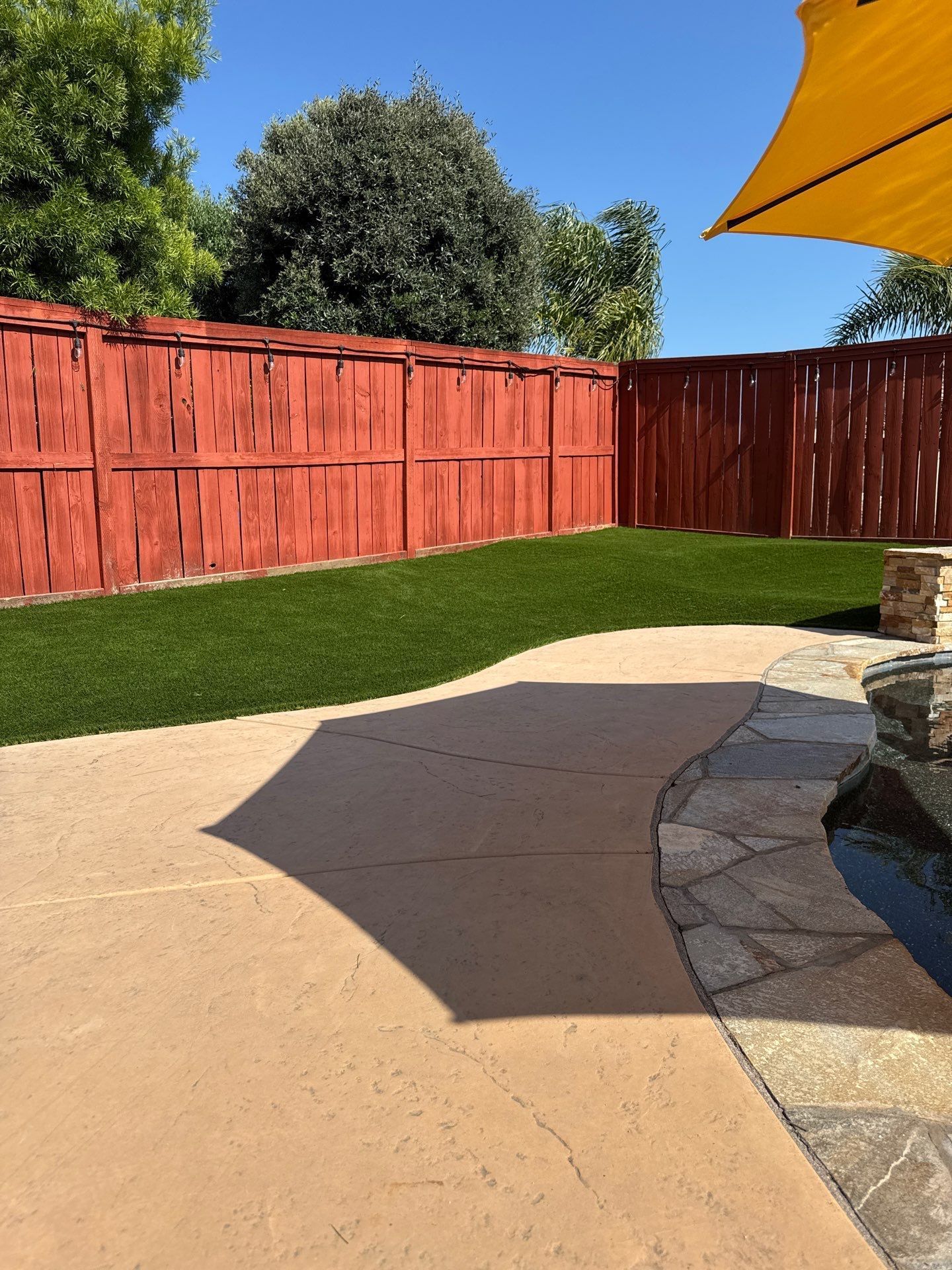 Backyard with red fence, green lawn, paved patio, and yellow umbrella.