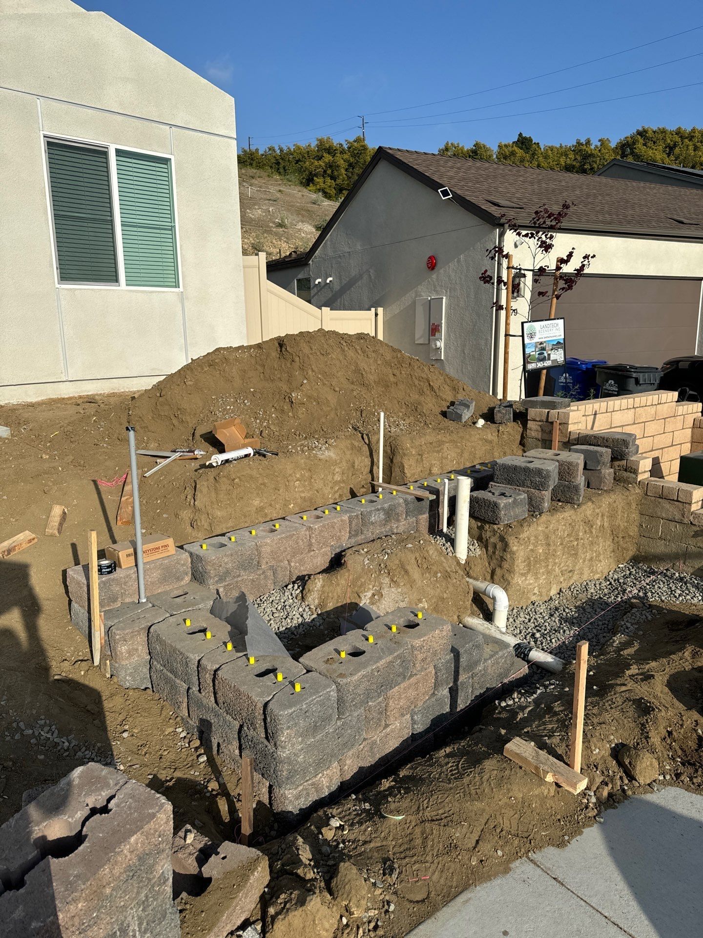 Construction of a retaining wall with grey blocks, dirt, and pipes, in front of a house under a blue sky.