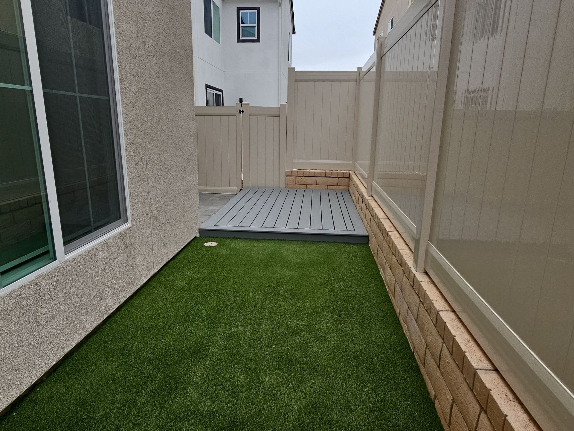 Small backyard with artificial turf, deck, beige fencing, and adjacent beige and brick wall.