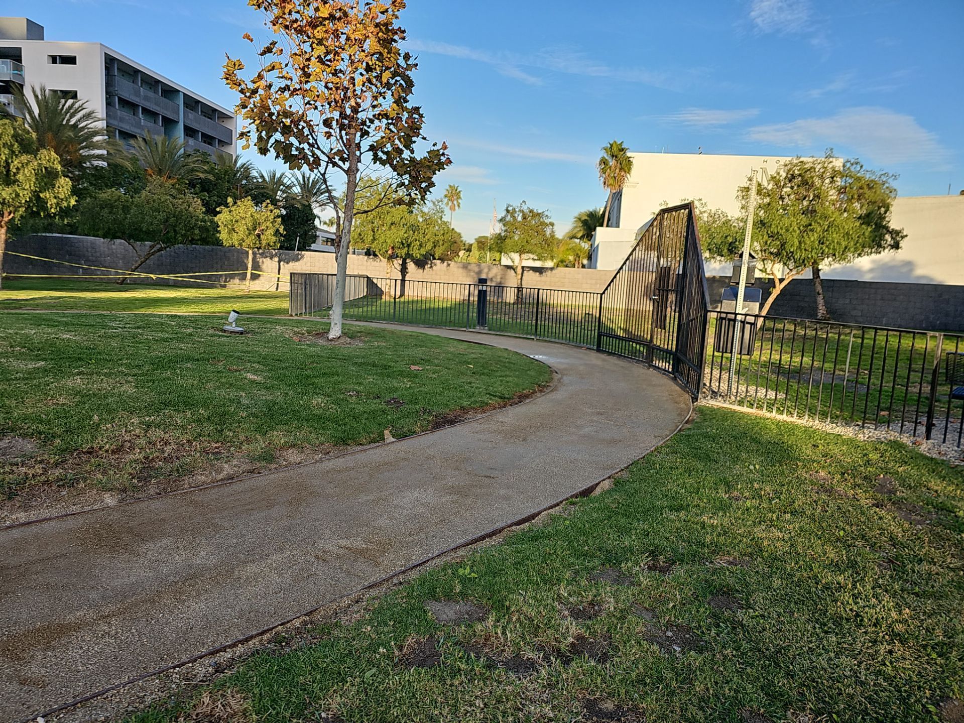 Gravel path winds through a park with green grass, trees, and a black fence. Buildings and blue sky in background.