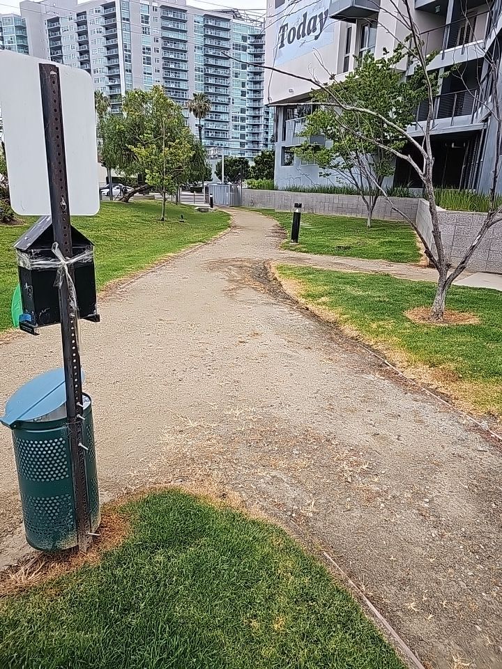 A gravel path through a park with grass, a trash can, and a tall building in the background.