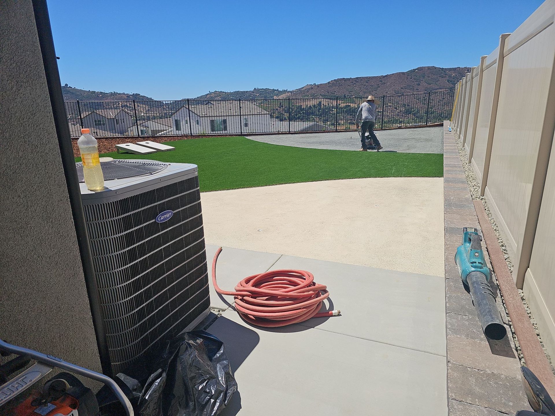 Air conditioner near a patio, hose, and a person walking in the background of a yard with artificial grass.
