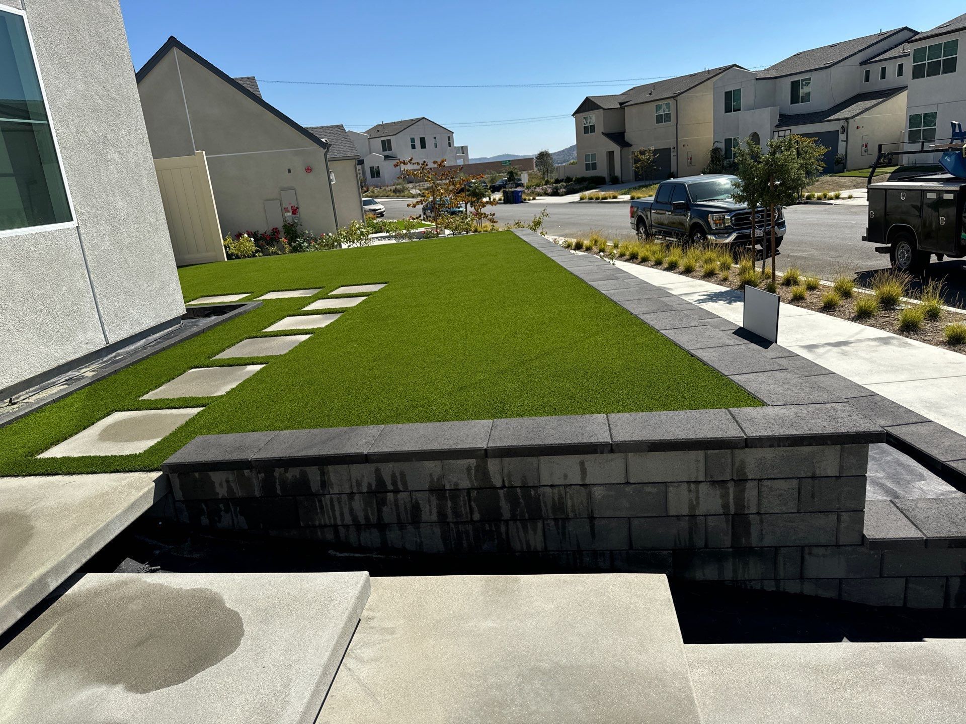 A front yard with artificial turf, stone retaining wall, and homes in the background.