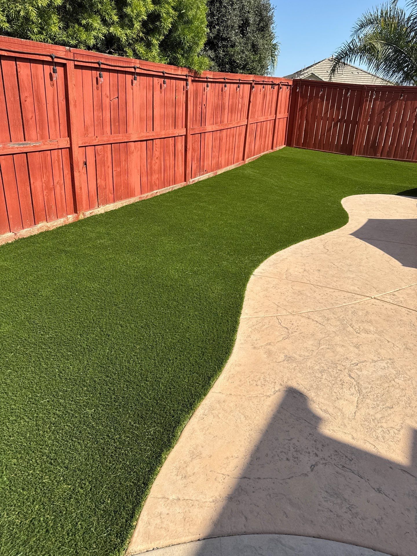 Green artificial turf next to a concrete patio and red wooden fence under a sunny sky.