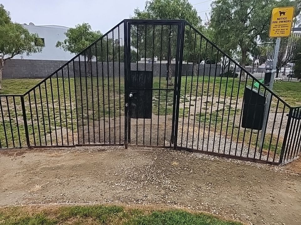 Black gated dog park with waste bag dispenser. Gravel and grass area.