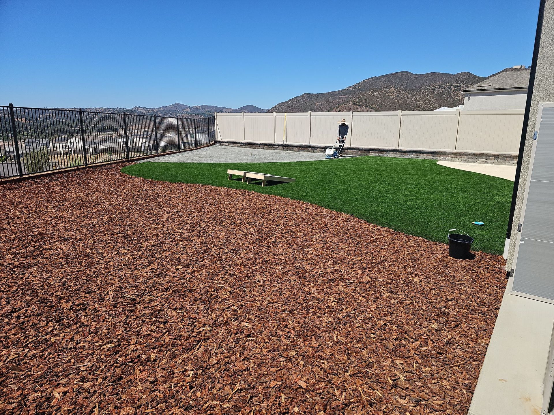 Backyard with wood chips, artificial turf, and fencing under a blue sky.