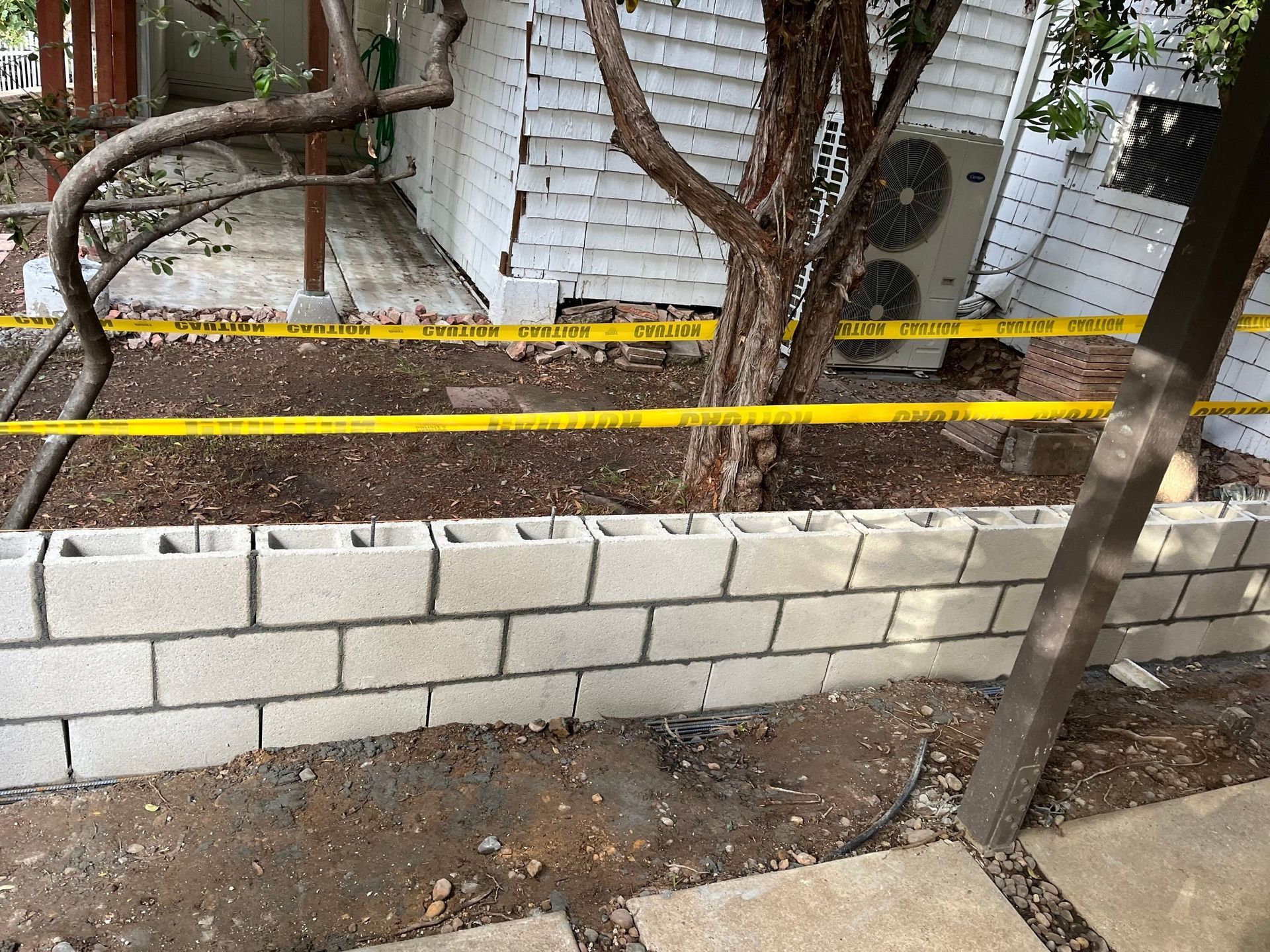 Cement block retaining wall with yellow caution tape, tree, and building in the background.