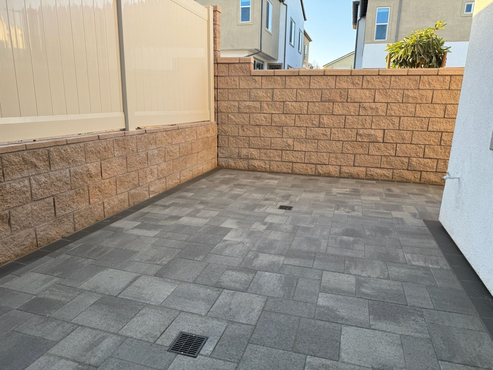 Enclosed paved patio with brown brick wall, beige fence, and a drainage grate.