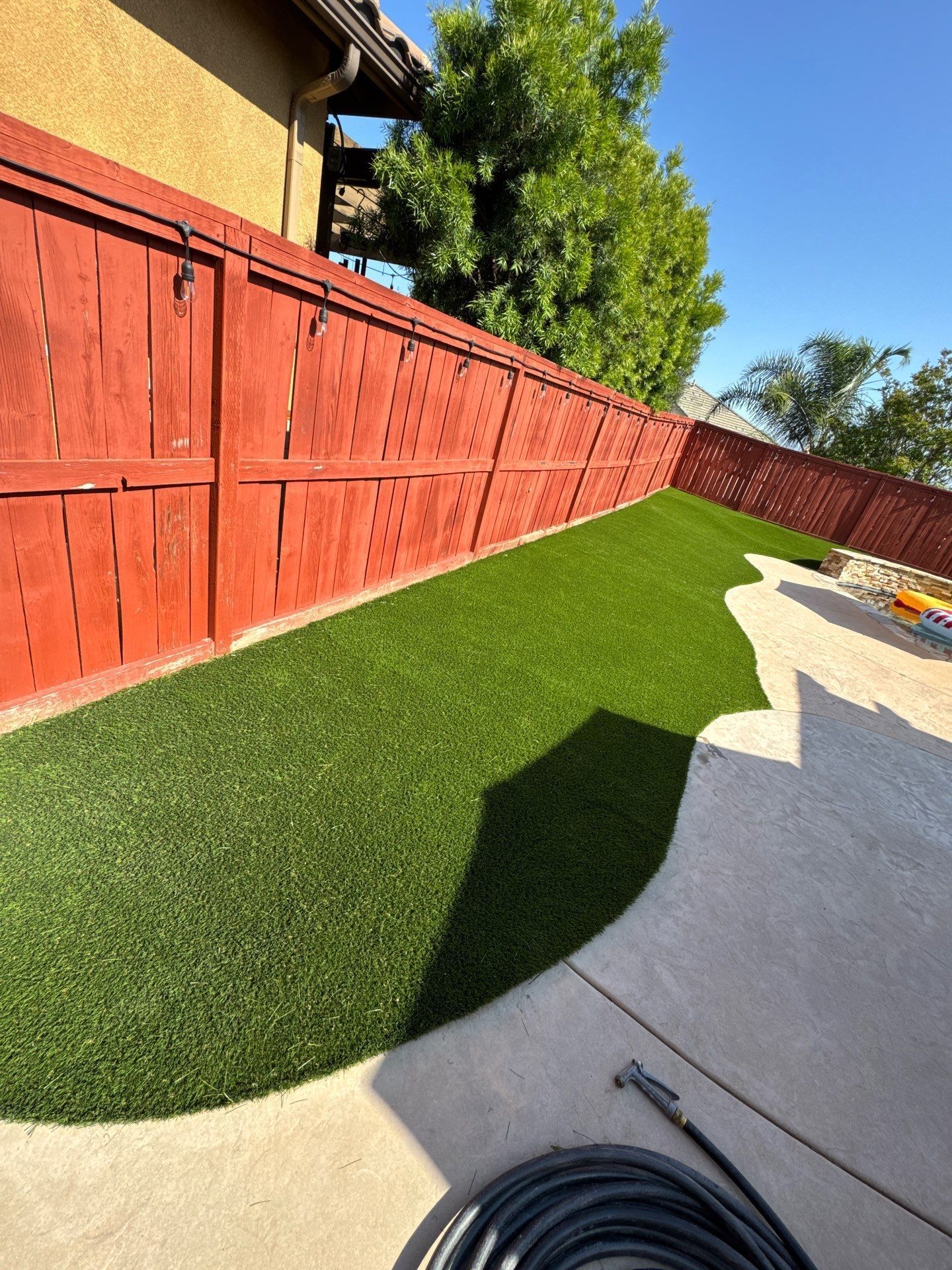 A backyard with red wooden fence, green artificial grass, and a concrete patio on a sunny day.