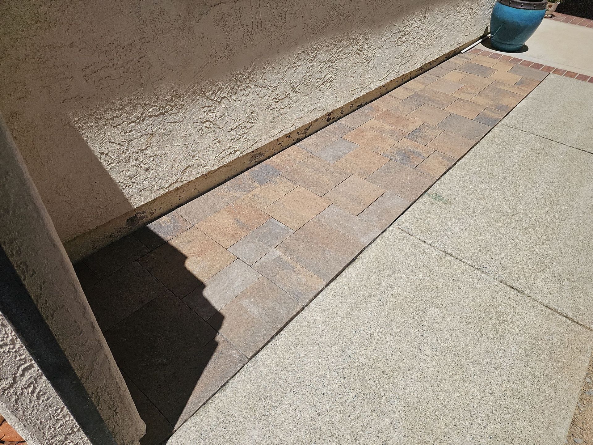 A narrow walkway of pavers next to a stucco wall, leading toward a potted plant.