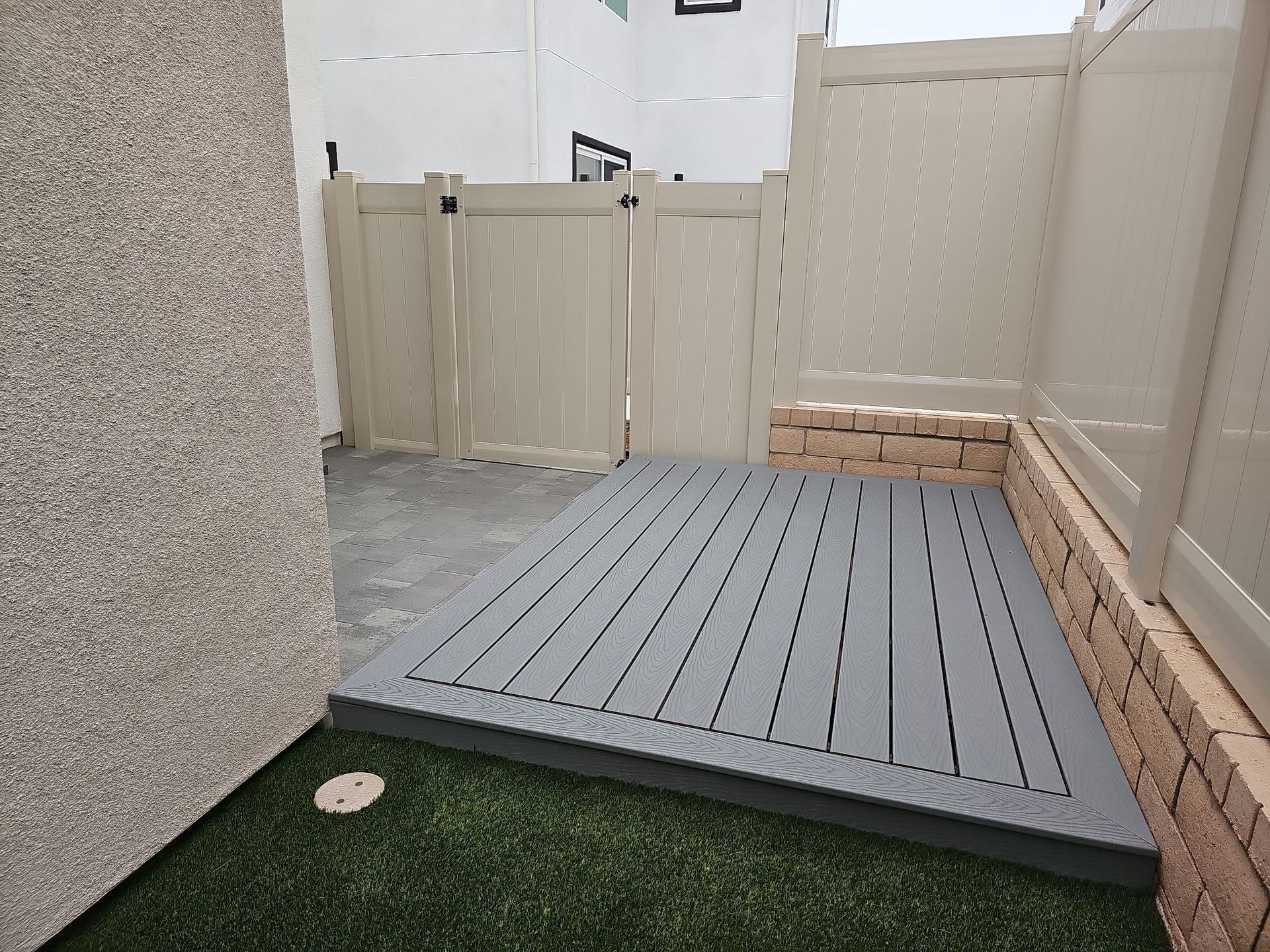 A small deck area with gray planks surrounded by beige fencing and brick.