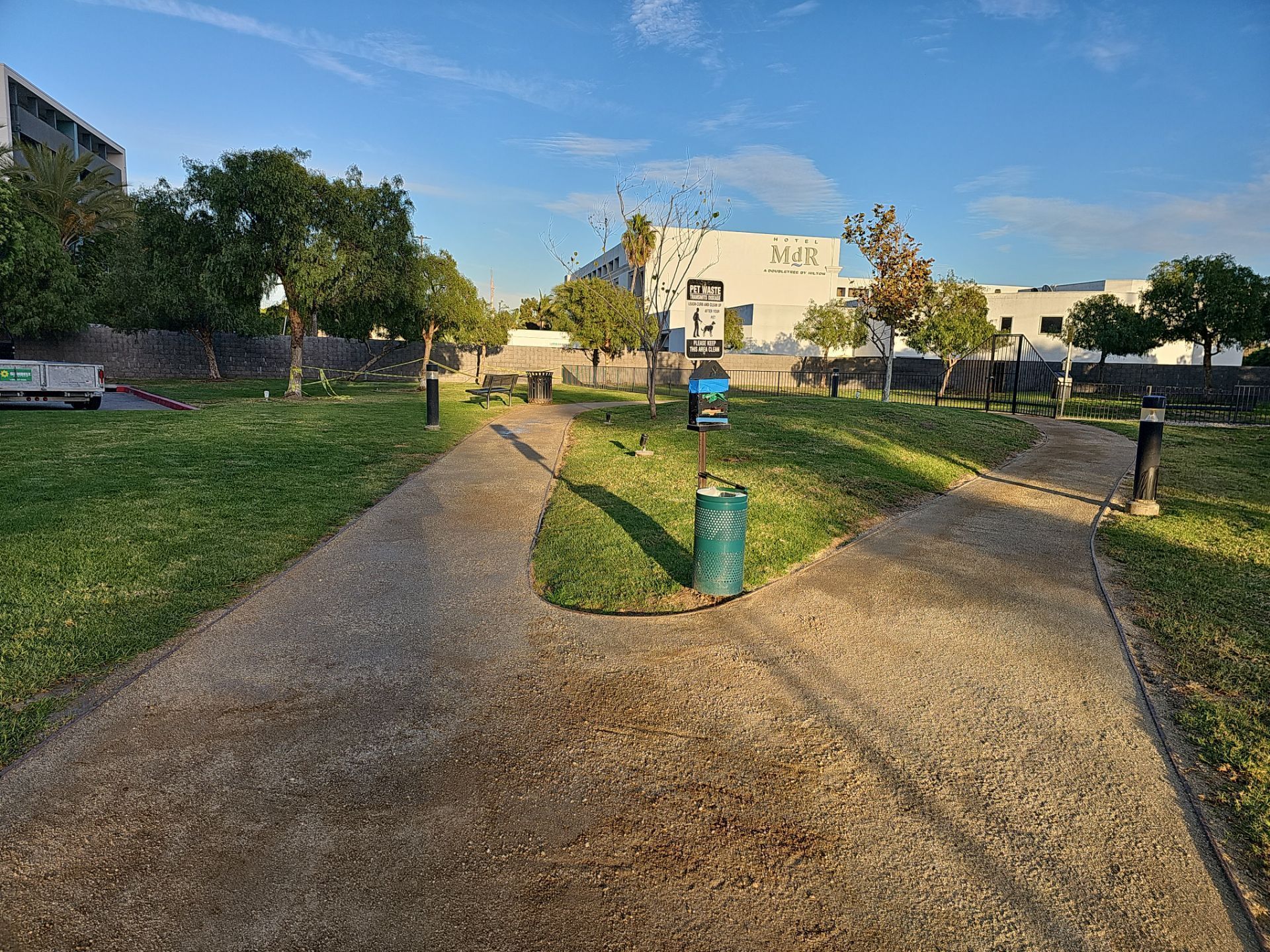 A park with a concrete path, green grass, trees, and buildings under a blue sky.