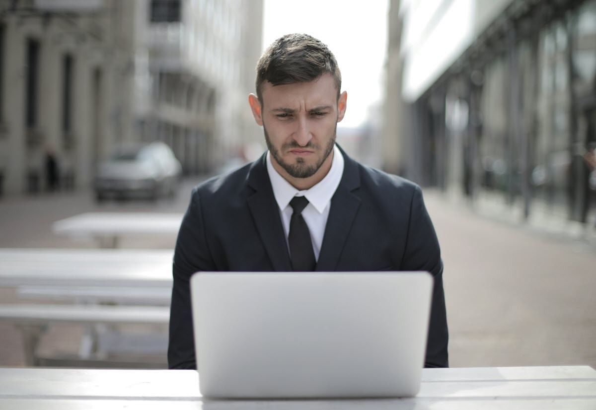 A person in a business suit with a furrowed expression works on a laptop at an outdoor table on a city street.