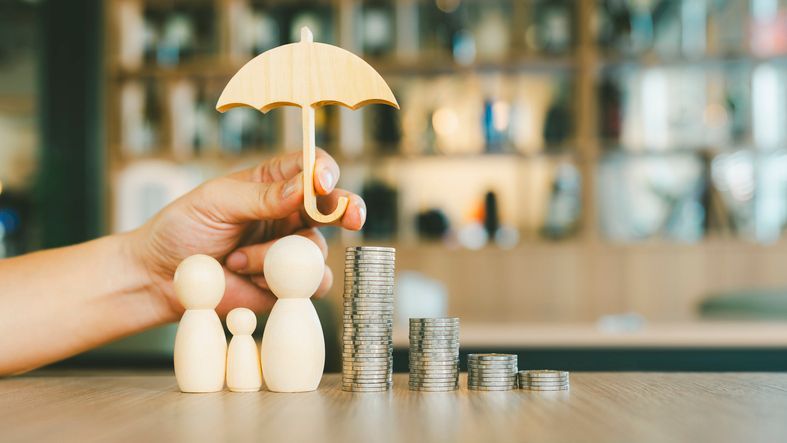 Hand holding wooden umbrella over wooden family figures and stacks of coins.