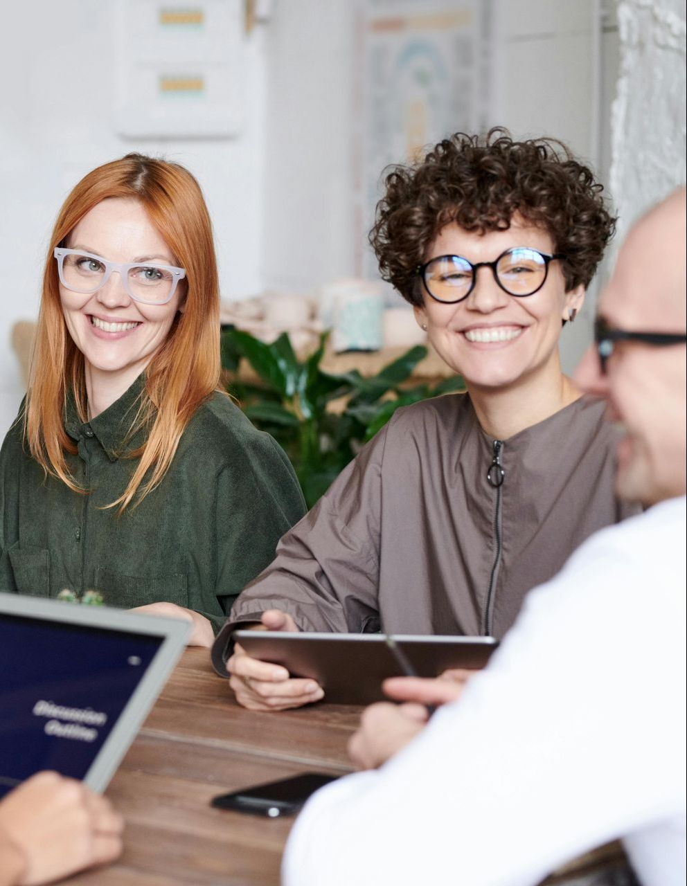 Three people smiling, sitting at a table with tablets. Plants and a blurred background are visible.